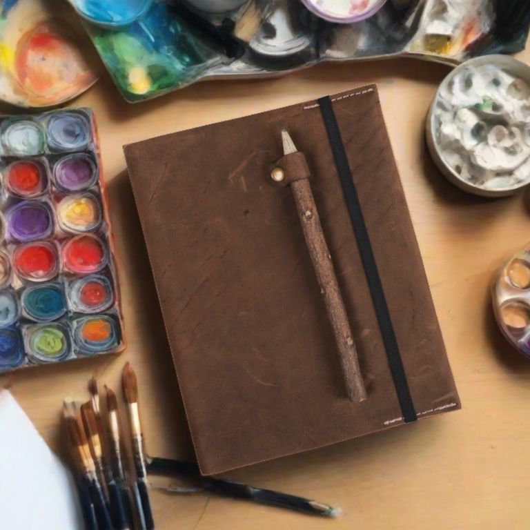 Brown leather notebook with a pen on a table surrounded by art supplies