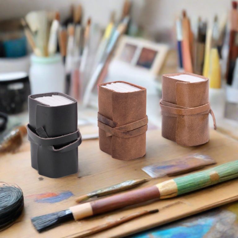 Three small cylindrical containers on a wooden surface with art supplies in the background