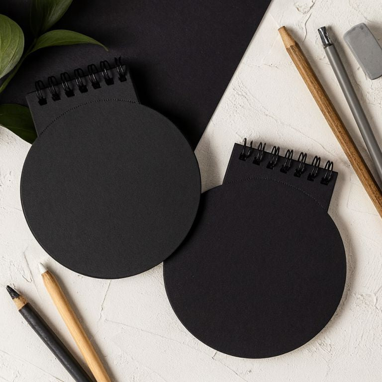 Two black circular notepads with spiral binding on a white surface with stationery items.
